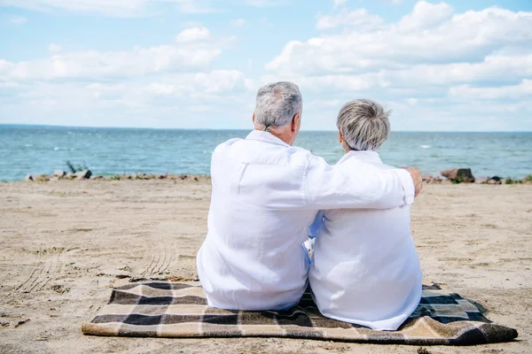 Задний вид пожилой пары в белых рубашках, сидящей на одеяле и обнимающейся на пляже — стоковое фото