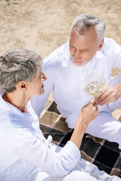 Vista aérea de pareja mayor sentada en manta y tintineo copas de vino con vino en la playa - foto de stock