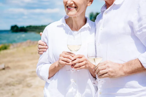 Vista ritagliata di sorridente coppia di anziani abbracciare e tenere bicchieri di vino con vino in spiaggia — Foto stock