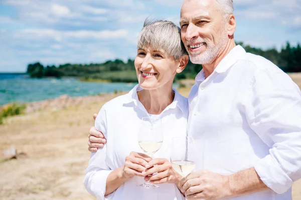 Sonriente pareja de ancianos abrazando y sosteniendo copas de vino con vino en la playa - foto de stock
