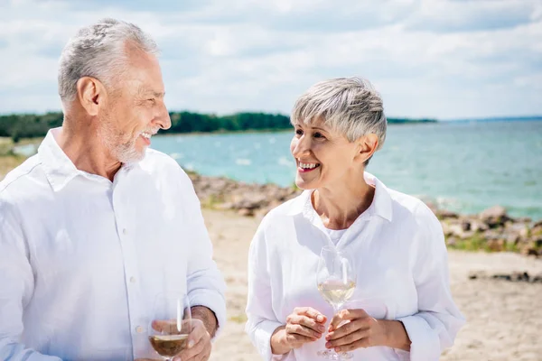 Sonriente pareja de ancianos sosteniendo copas de vino con vino y mirándose en la playa - foto de stock