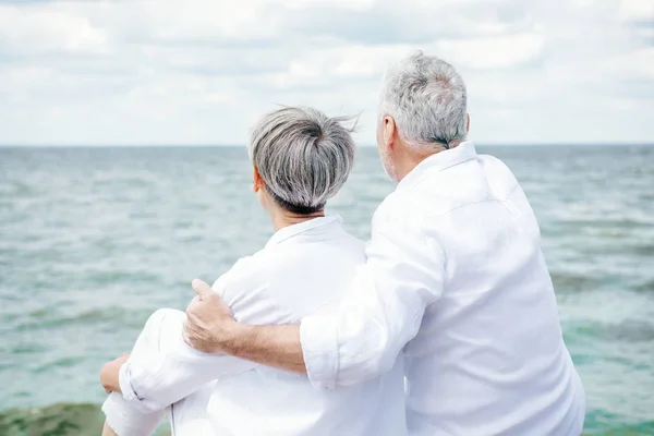 Back view of senior couple in white shirts embracing near river under blue sky — Stock Photo