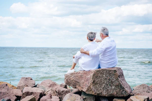 Vista trasera de pareja mayor en camisas blancas abrazando cerca del río bajo el cielo azul - foto de stock