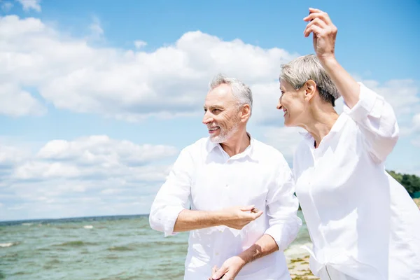 Щаслива усміхнена старша пара в білих сорочках жестикулює на пляжі під блакитним небом — стокове фото