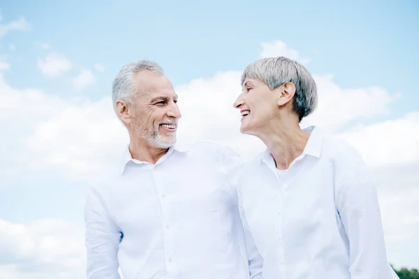 Feliz sorrindo casal sênior em camisas brancas olhando um para o outro sob o céu azul — Fotografia de Stock
