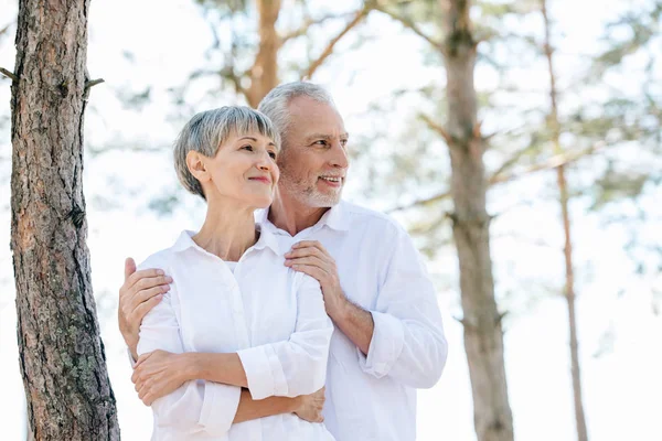 Sonriente pareja de ancianos en camisas blancas abrazando y mirando hacia otro lado en el bosque - foto de stock