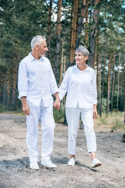Full length view of happy senior couple in white shirts holding hands and looking at each other in forest — Stock Photo