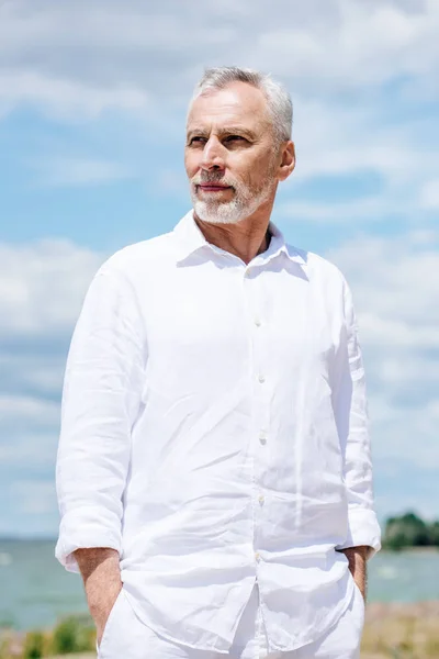 Pensive senior man in white shirt standing with hands in pockets under blue sky — Stock Photo