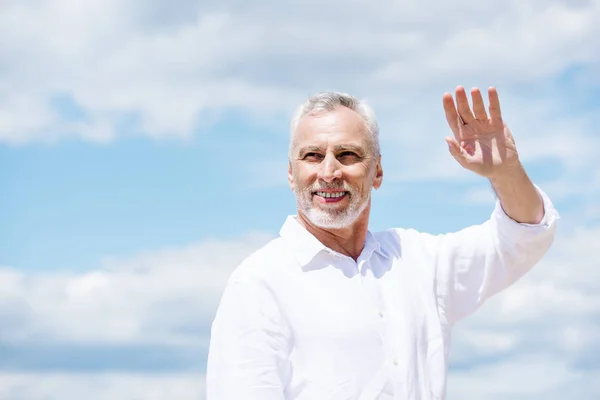 Uomo anziano sorridente con la barba distogliendo lo sguardo e agitando la mano sotto il cielo blu nella giornata di sole — Foto stock