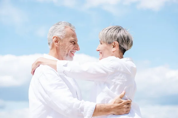 Seitenansicht eines lächelnden Seniorenpaares, das sich unter blauem Himmel umarmt und ansieht — Stockfoto