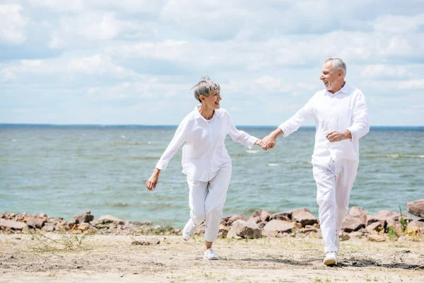 Vista completa de la pareja de ancianos tomados de la mano y mirándose en la playa - foto de stock