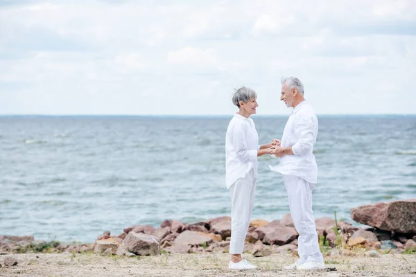 Ganzkörperansicht eines älteren Paares, das sich am Strand an den Händen hält und einander ansieht — Stockfoto