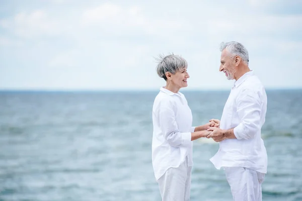 Vista lateral de feliz pareja de ancianos tomados de la mano y mirándose el uno al otro cerca del río - foto de stock