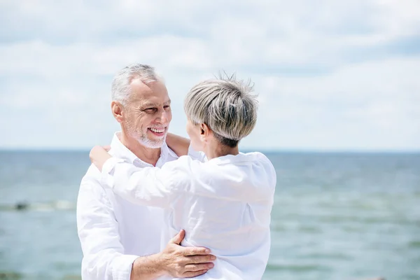 Feliz pareja de ancianos en camisas blancas abrazándose y mirándose bajo el cielo azul - foto de stock