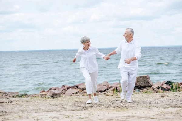 Pleine longueur vue de heureux couple de personnes âgées tenant la main et se regardant tout en courant à la plage — Photo de stock