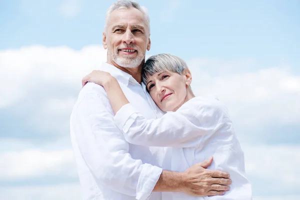 Feliz casal sênior em camisas brancas abraçando sob o céu azul — Fotografia de Stock