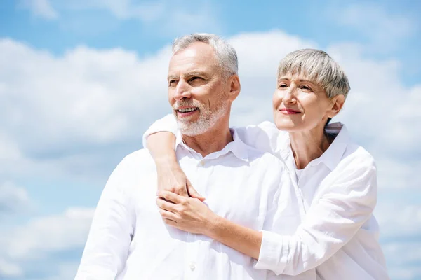 Feliz pareja de ancianos en camisas blancas abrazándose bajo el cielo azul - foto de stock