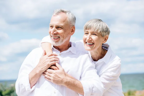 Feliz casal sênior em camisas brancas abraçando sob o céu azul — Fotografia de Stock