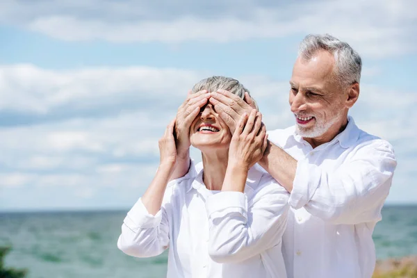 Улыбающийся пожилой мужчина закрывает глаза на жену возле реки в солнечный день — стоковое фото