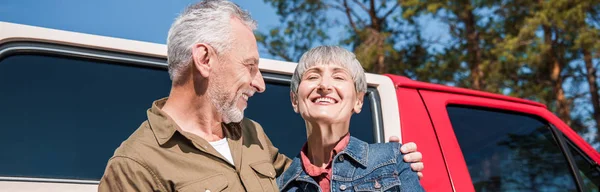Панорамний вид на щасливу старшу пару туристів, що стоять біля машини і обіймаються з посмішкою — стокове фото