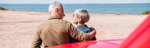 Vista panorámica de la pareja de turistas mayores de pie cerca de coche y abrazo en la playa - foto de stock