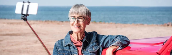 Vista panoramica della donna anziana sorridente in piedi vicino alla macchina e scattare selfie in spiaggia — Foto stock