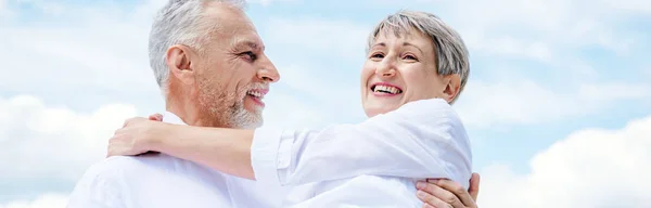 Panoramablick auf einen lächelnden Senior, der seine Frau unter blauem Himmel abhebt — Stockfoto