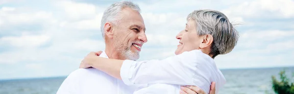 Панорамный вид на улыбающегося пожилого мужчину, поднимающего жену под голубым небом — стоковое фото