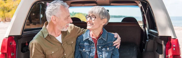 Vista panorámica de sonriente pareja de ancianos abrazándose y mirándose el uno al otro cerca del coche - foto de stock