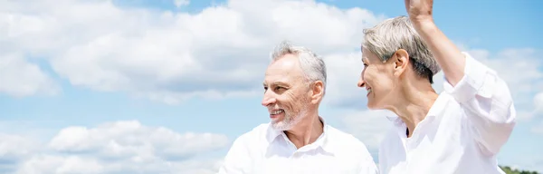 Vista panorámica de la sonriente pareja de ancianos en camisas blancas mirando hacia otro lado bajo el cielo azul - foto de stock