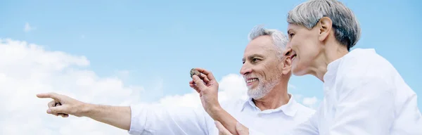 Vista panorámica de la sonriente pareja de ancianos en camisas blancas mirando hacia otro lado y señalando con el dedo bajo el cielo azul - foto de stock