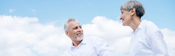 Панорамный вид улыбающейся пожилой пары в белых рубашках, смотрящей друг на друга под голубым небом — стоковое фото