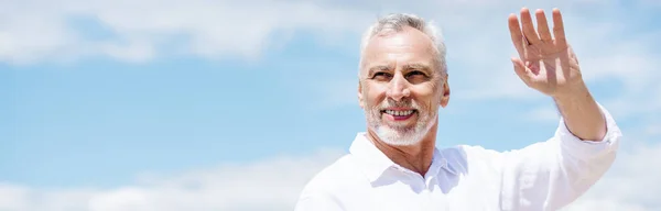 Panoramablick auf einen lächelnden Senior im weißen Hemd, der unter blauem Himmel mit der Hand wedelt — Stockfoto