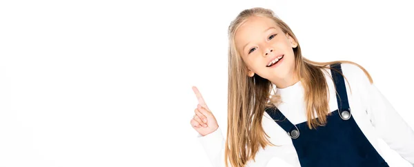 Panoramic shot of cheerful schoolgirl pointing with finger and smiling at camera isolated on white — Stock Photo