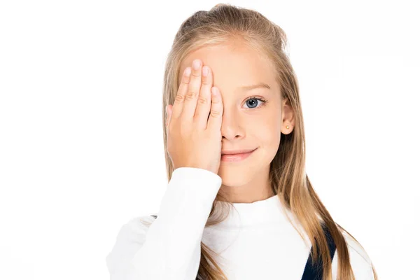 Entzückendes, fröhliches Schulmädchen, das Auge mit der Hand bedeckt, während es isoliert auf weiß in die Kamera schaut — Stockfoto