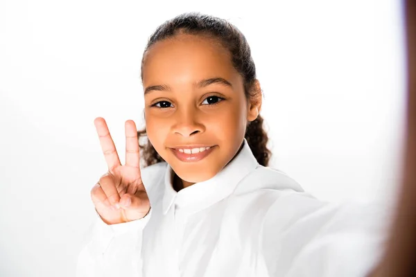 Enfoque selectivo de alegre afroamericana colegiala mostrando signo de victoria sobre fondo blanco - foto de stock