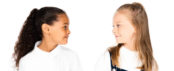 Plano panorámico de dos colegialas sonrientes multiculturales mirándose aisladas en blanco - foto de stock