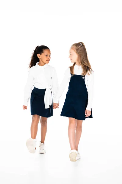 Вигляд на повну довжину двох мультикультурних школярок, що тримаються за руки і йдуть разом на білому тлі — стокове фото