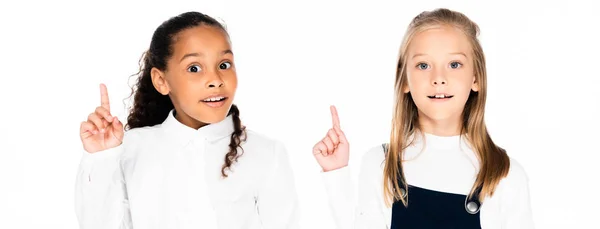 Tiro panorâmico de duas colegiais multiculturais bonitos olhando para a câmera e mostrando gestos ideia isolado no branco — Fotografia de Stock