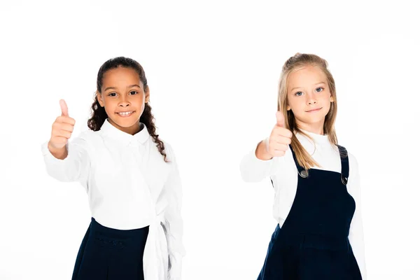 Dos sonrientes colegialas multiculturales mostrando los pulgares hacia arriba mientras mira a la cámara aislada en blanco - foto de stock