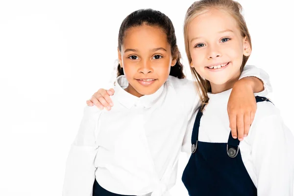 Deux écolières multiculturelles heureux étreignant et souriant à la caméra isolé sur blanc — Photo de stock