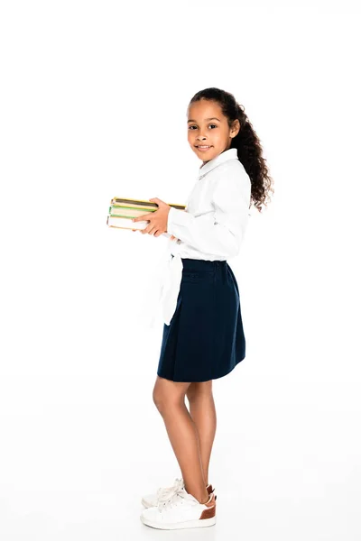 Повнометражний вид на чарівну афроамериканську школярку, яка тримає книги і посміхається на камеру на білому тлі — стокове фото