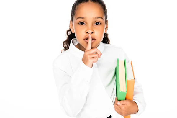 Cute african american schoolgirl showing hush gesture while holding books isolated on white — Stock Photo
