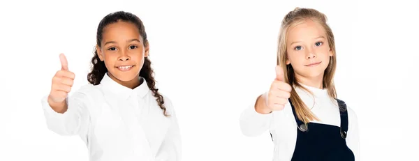 Plano panorámico de dos colegialas multiculturales mostrando el pulgar hacia arriba mientras mira la cámara aislada en blanco - foto de stock