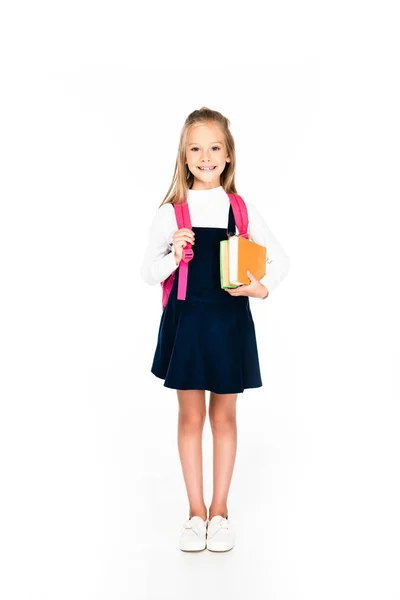 Vista de longitud completa de linda colegiala sonriendo a la cámara mientras sostiene libros sobre fondo blanco — Stock Photo