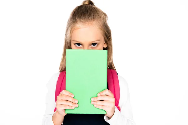 Carino studentessa guardando fotocamera mentre oscura il viso con libro isolato su bianco — Foto stock