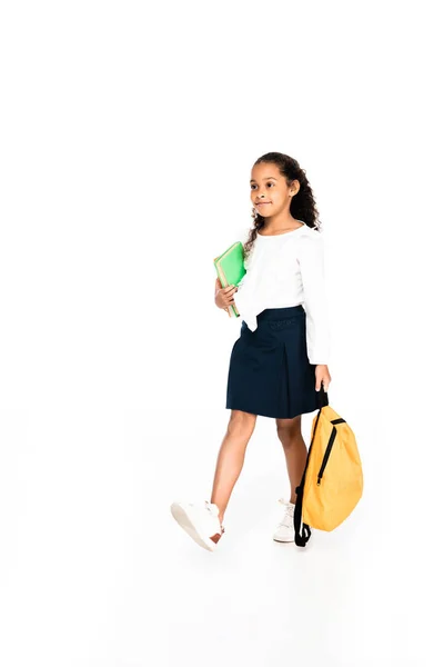 Full length view of African American schoolgirl walking with backpack and book on white background — Fotografia de Stock