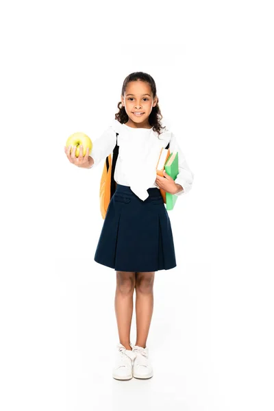 Vue pleine longueur de l'adorable écolière afro-américaine souriant à la caméra tout en tenant des livres et de la pomme sur fond blanc — Photo de stock