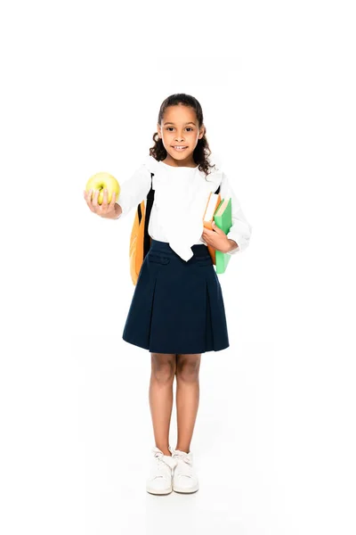 Vista completa de la adorable colegiala afroamericana sonriendo a la cámara mientras sostiene libros y manzana sobre fondo blanco - foto de stock