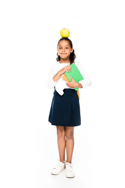 Full length view of cheerful African american schoolgirl with apple on head holding books on white background — стоковое фото